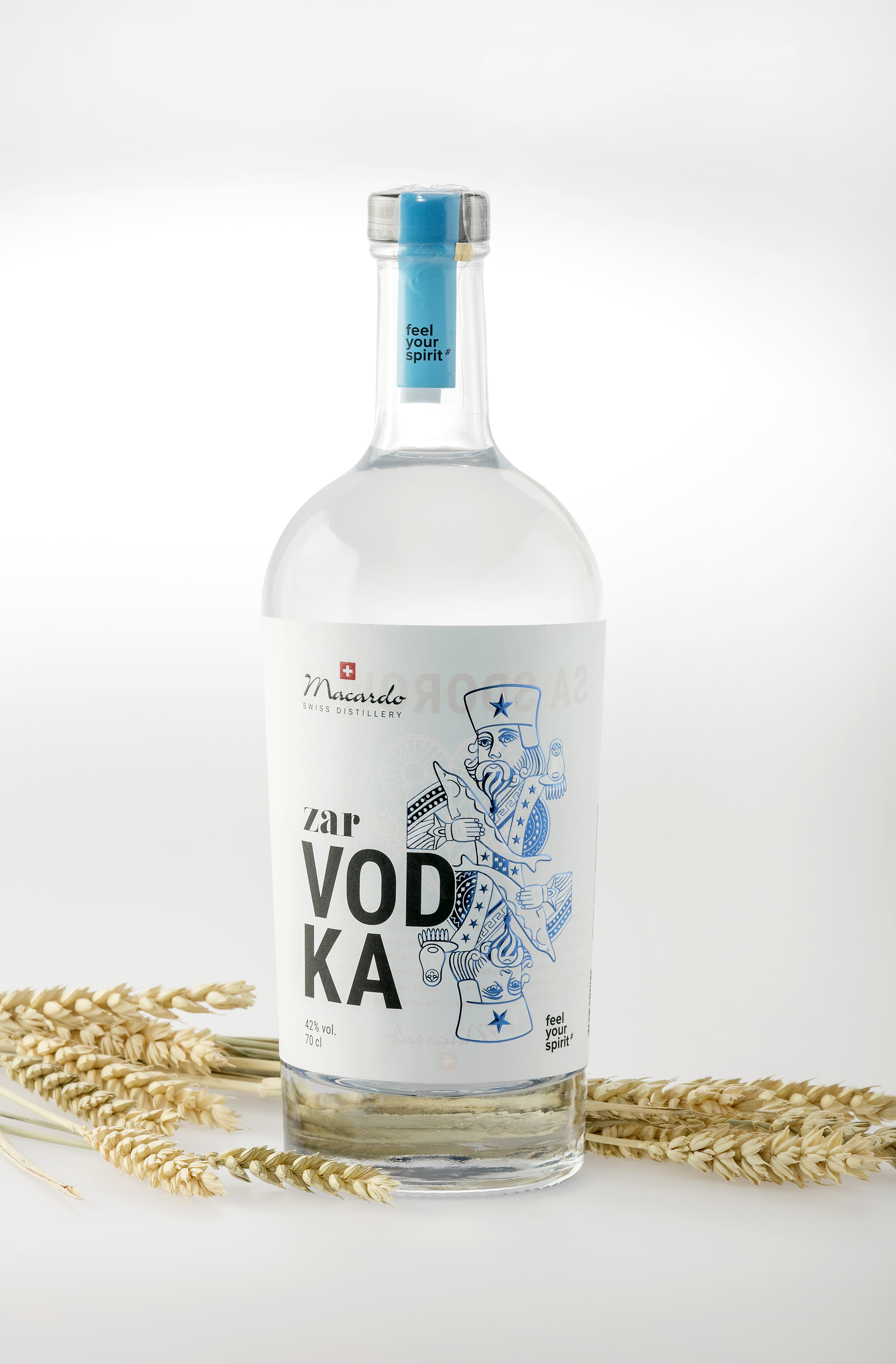 Eine klare Glasflasche Zar Vodka der Macardo Swiss Distillery steht vor hellem Hintergrund. Das Etikett zeigt den Schriftzug "Zar Vodka" und eine blaue Illustration. Goldene Weizenähren liegen dekorativ um die Flasche. Der Verschluss ist blau.